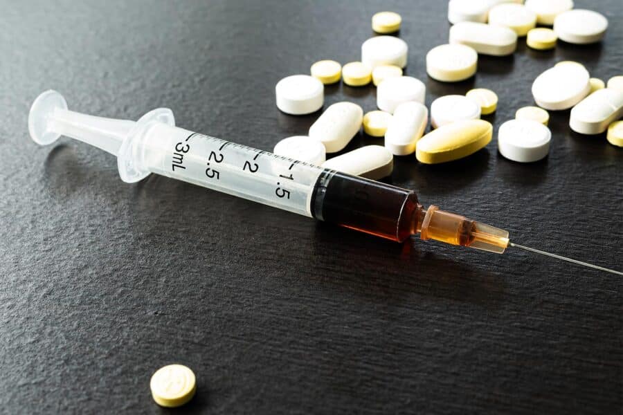 Gateway Drugs: A dark surface with a syringe containing a dark liquid and several different types of pills.